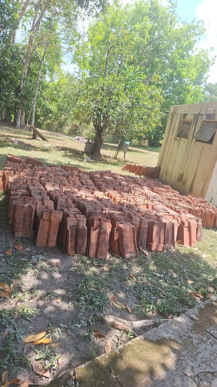 Centenas de telhas cerâmicas empilhadas sobre a grama do terreno, ao lado de uma construção existente